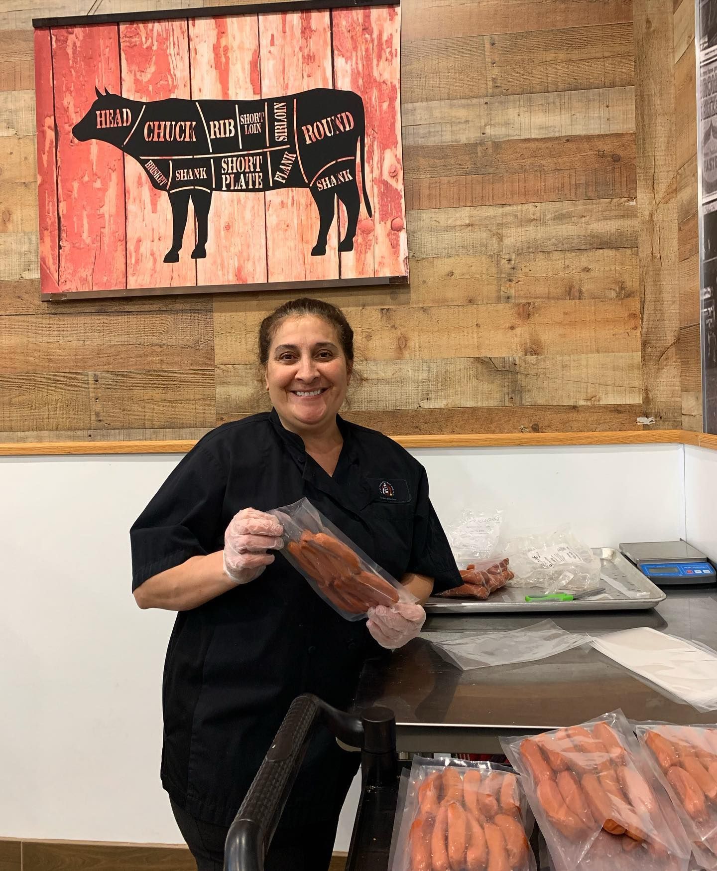Woman in black holding packaged sausages, smiling in a butcher shop, cow diagram on the wall.