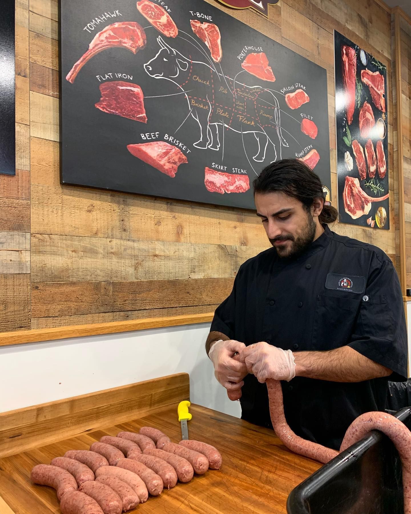 Butcher in black uniform making sausages at a wooden counter, with a diagram of meat cuts on the wall.
