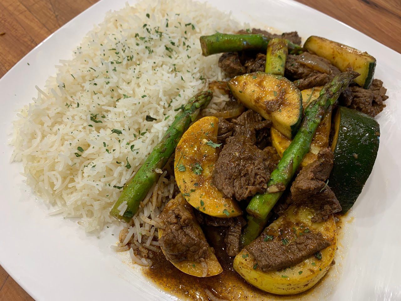 Plate of white rice with beef, zucchini, and asparagus in a dark sauce, garnished with herbs.
