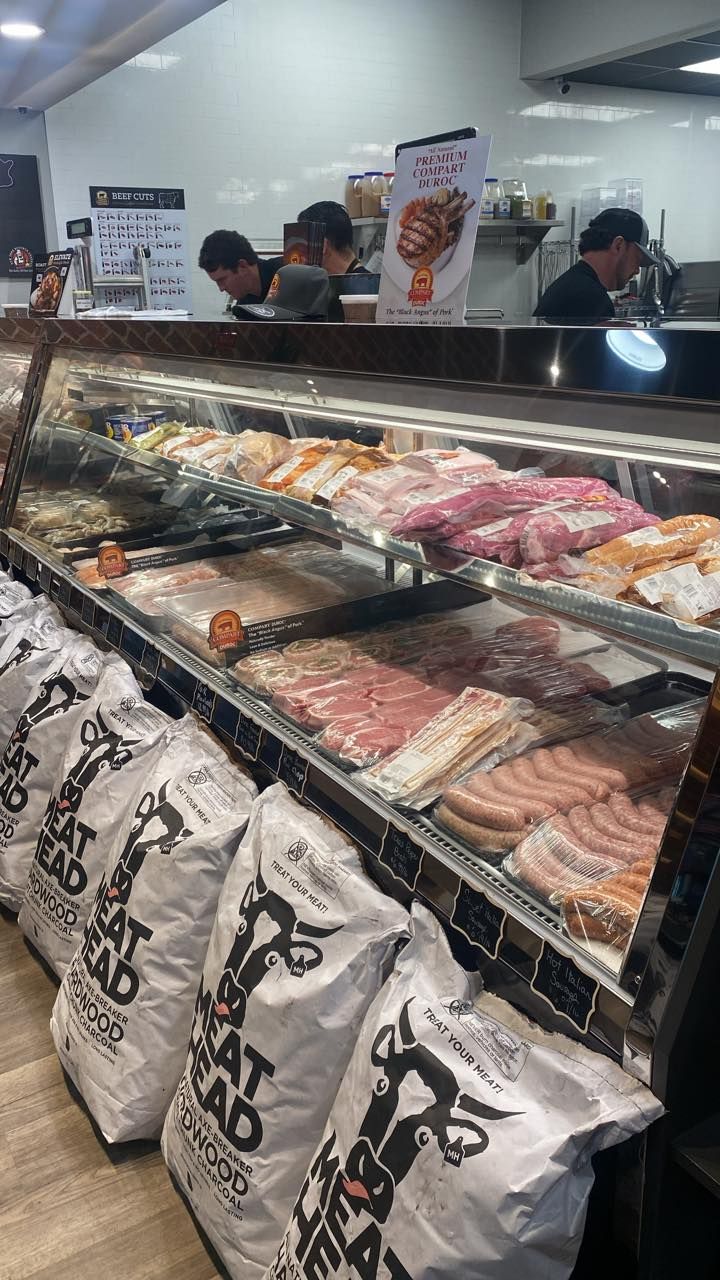 Meat display case with various cuts and sausages, bags of charcoal, workers behind counter.