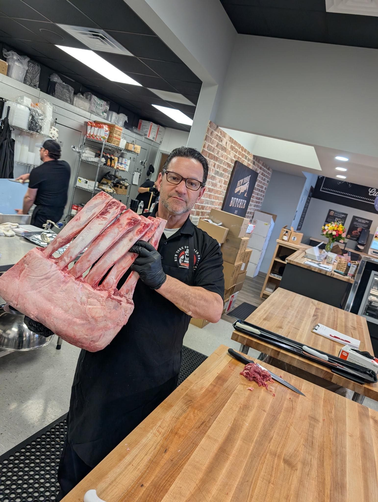 Butcher holds a large, raw rib roast in a butcher shop. He wears black gloves, a black apron and has a focused expression.