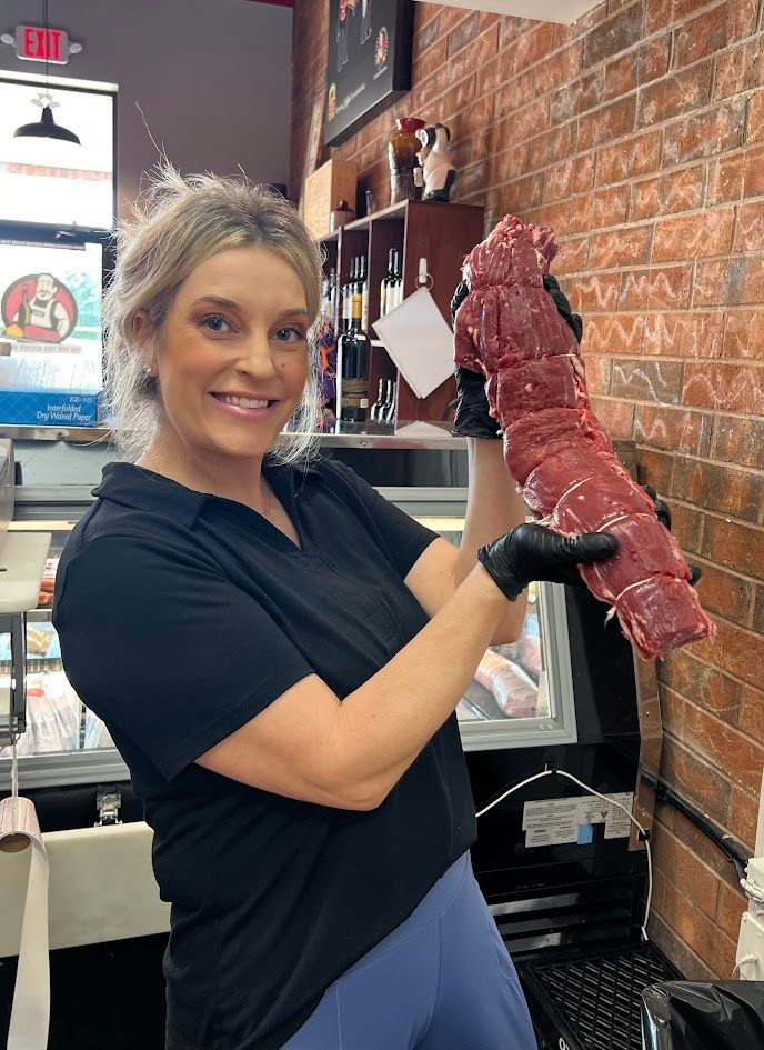 Woman in black holding a large cut of red meat in a butcher shop, smiling.