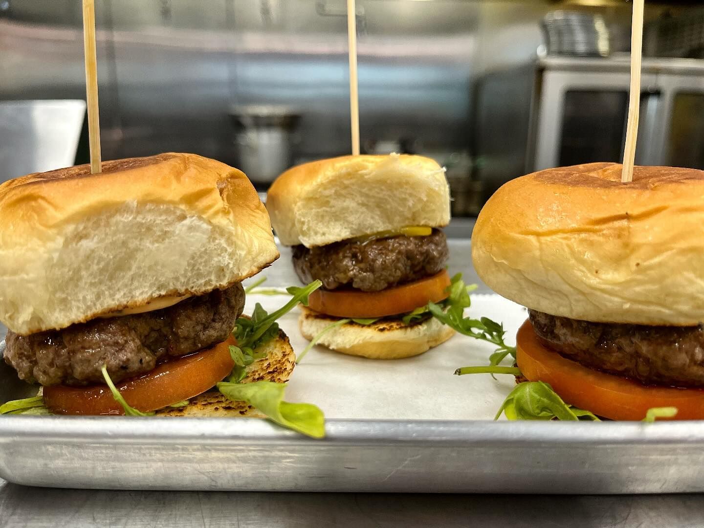 Three sliders on a tray: beef patties on buns, tomato slices, arugula, skewers.