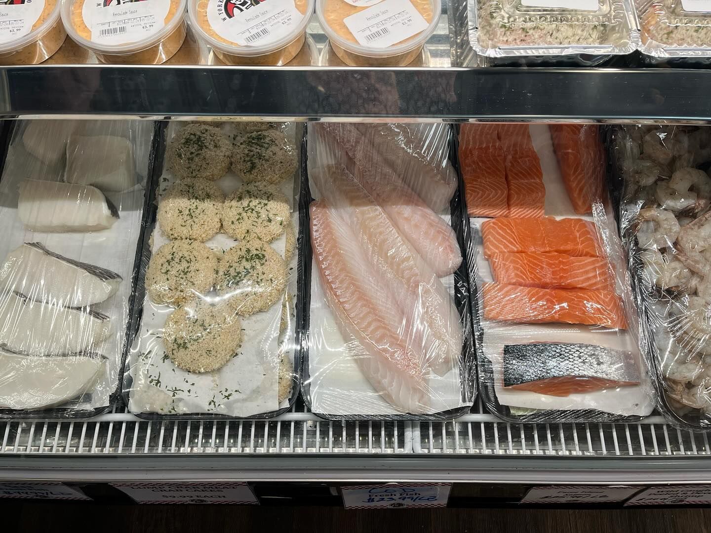 Display case with various prepared fish: salmon, tilapia, and fish cakes, covered in plastic wrap.