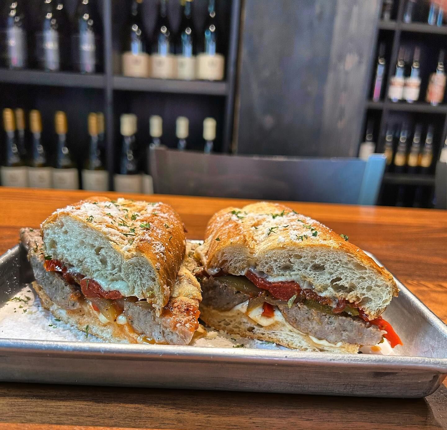 Sausage sandwich cut in half on a metal tray. Red peppers, and cheese between bread; wine bottles in the background.