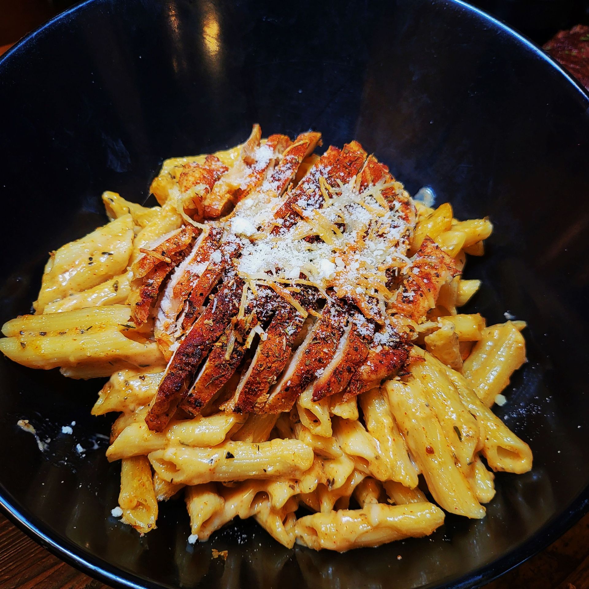 Penne pasta with sliced, grilled chicken in a dark bowl, topped with grated parmesan cheese.