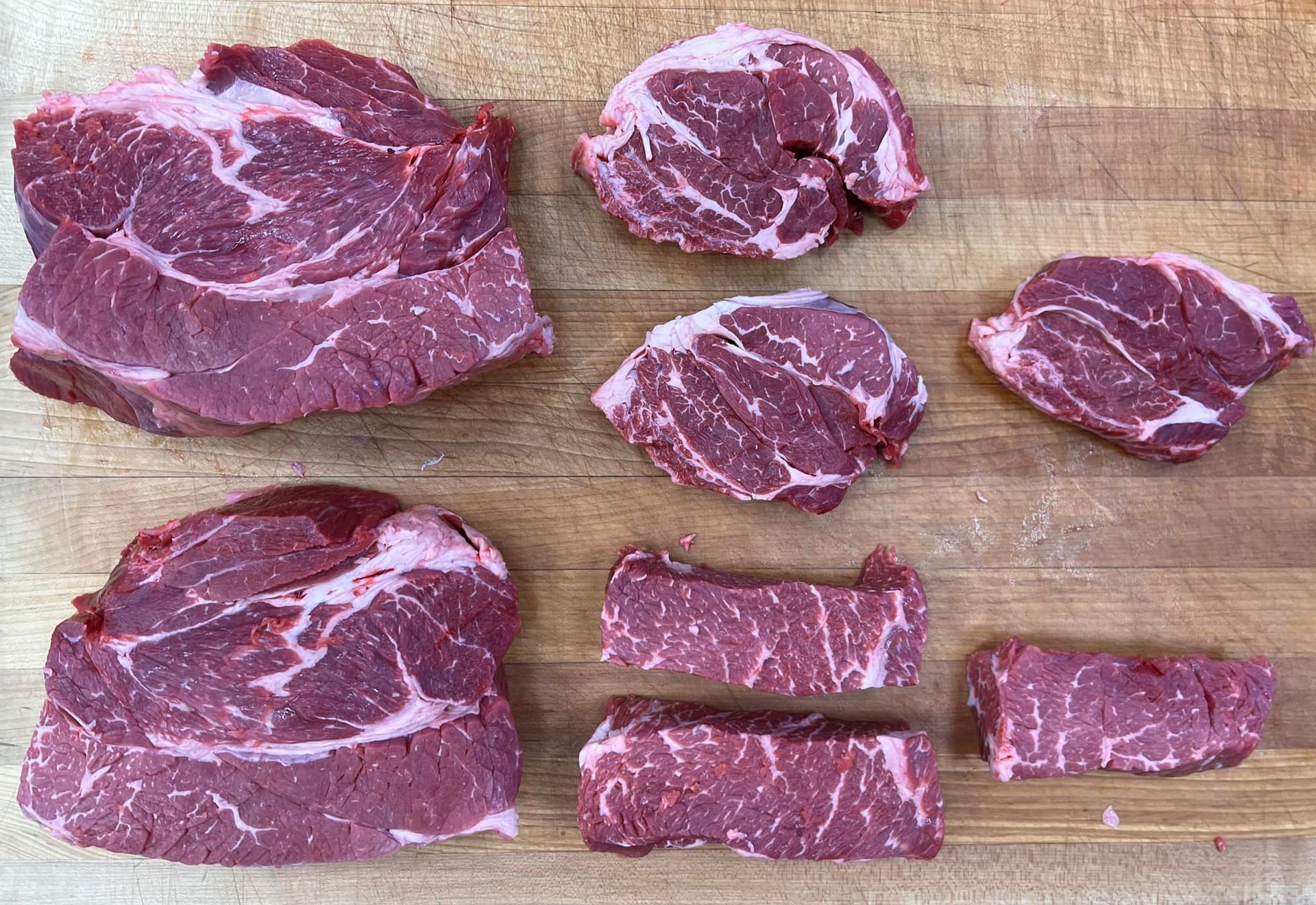Raw steaks on a wooden cutting board; various cuts and sizes, red meat with white marbling.