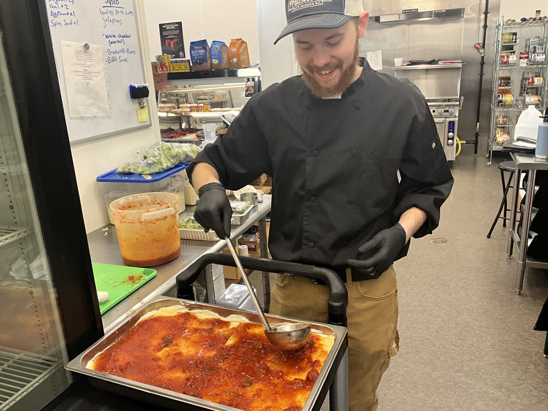 Chef ladles sauce over a lasagna in a commercial kitchen; he smiles.
