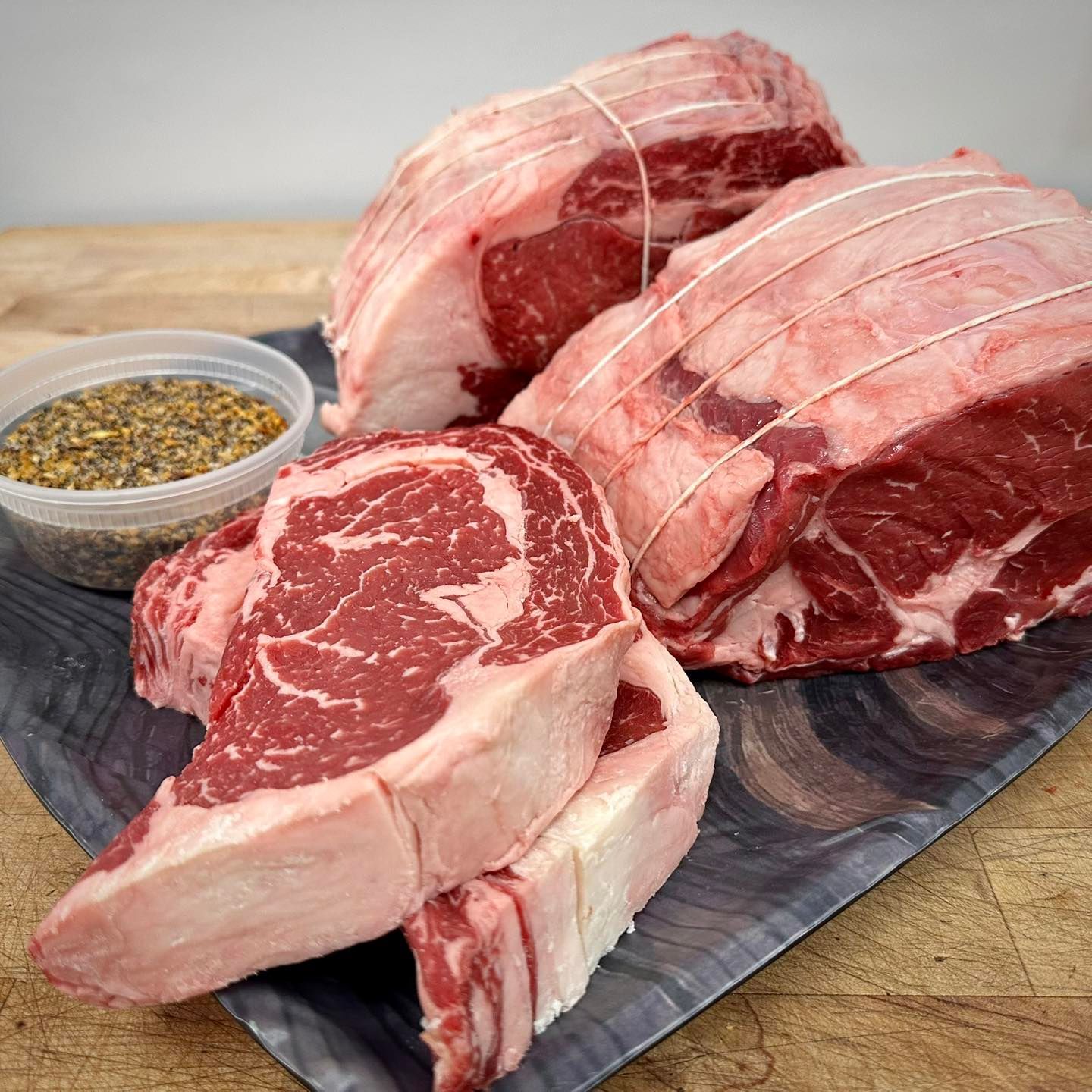 Raw ribeye steaks, roast beef tied with twine, and seasoning on a marble tray.