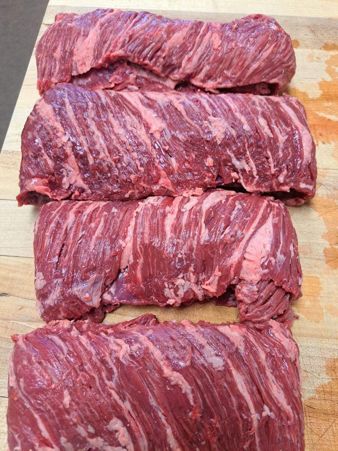 Four raw, red flank steaks arranged on a wooden surface, showing visible marbling.