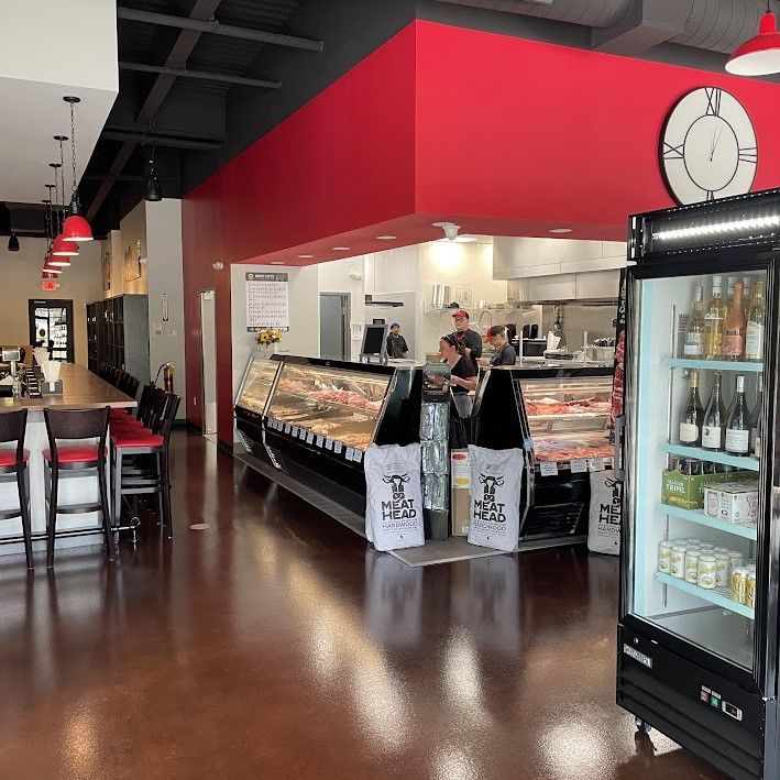 Interior of a deli with red accents, a refrigerated drinks case, and customers near the counter.