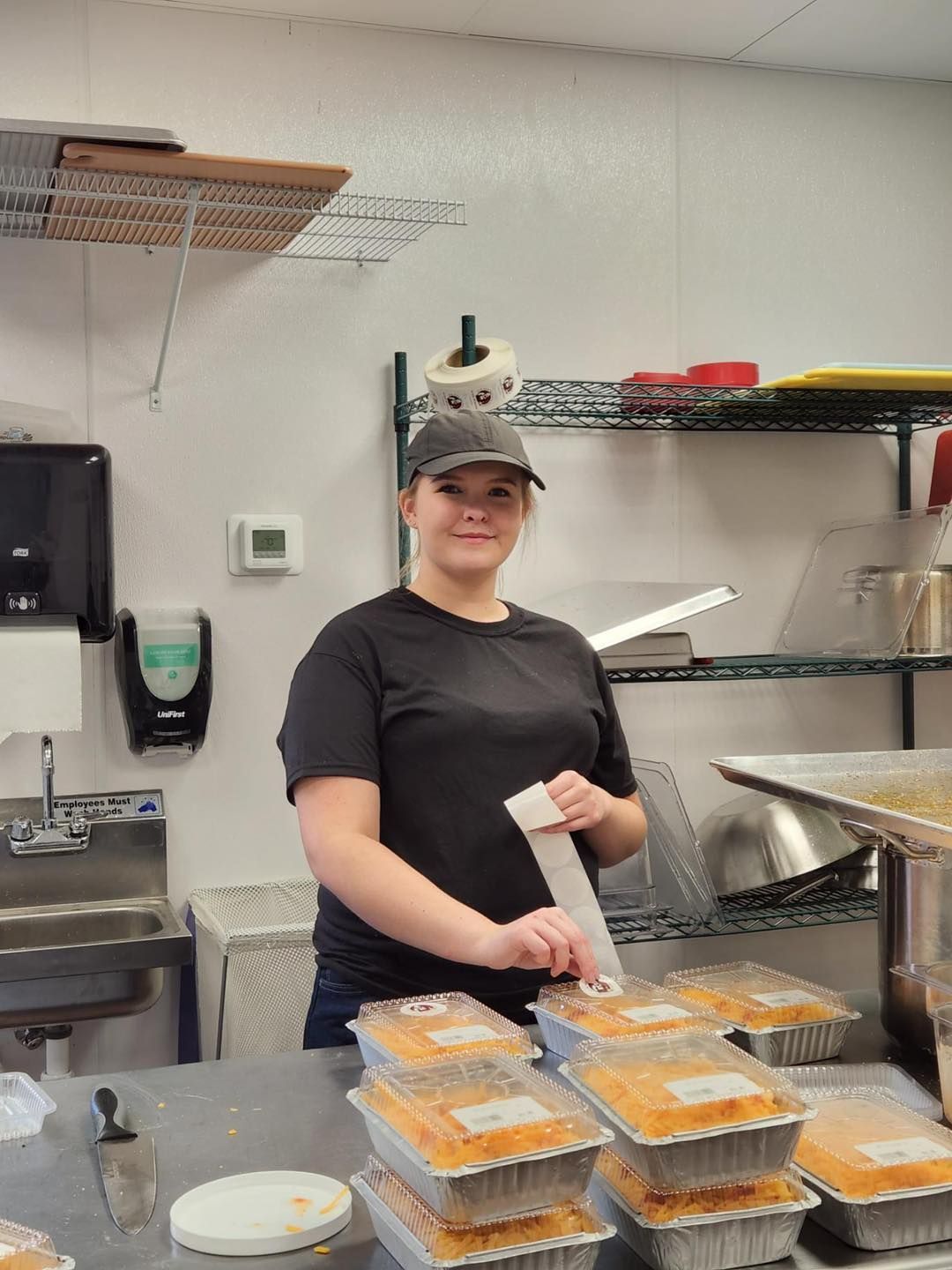 Woman in kitchen, packaging food, wearing a cap and smiling, near stacked containers.