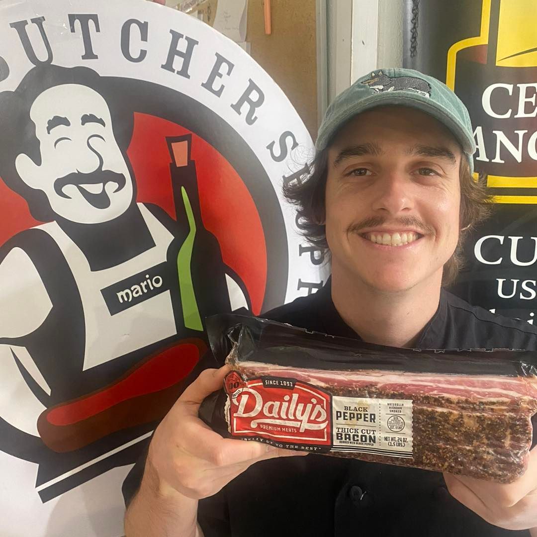Man holding a package of Daily's peppered bacon in a butcher shop, smiling. Behind him is a logo.