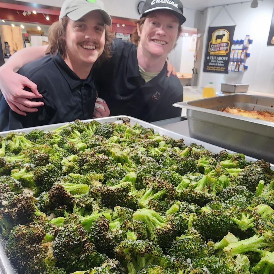 Two smiling men with arms around each other behind a tray of roasted broccoli.