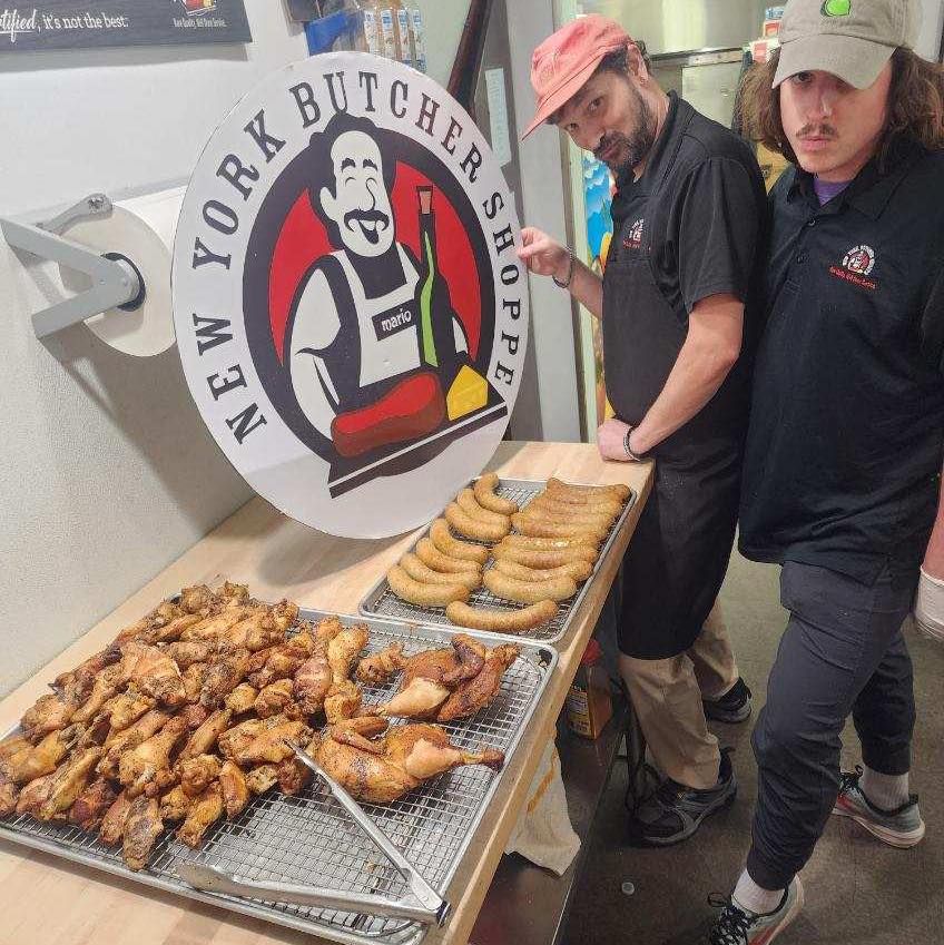 Two men stand near a New York Butcher Shoppe sign with prepared food. One points.