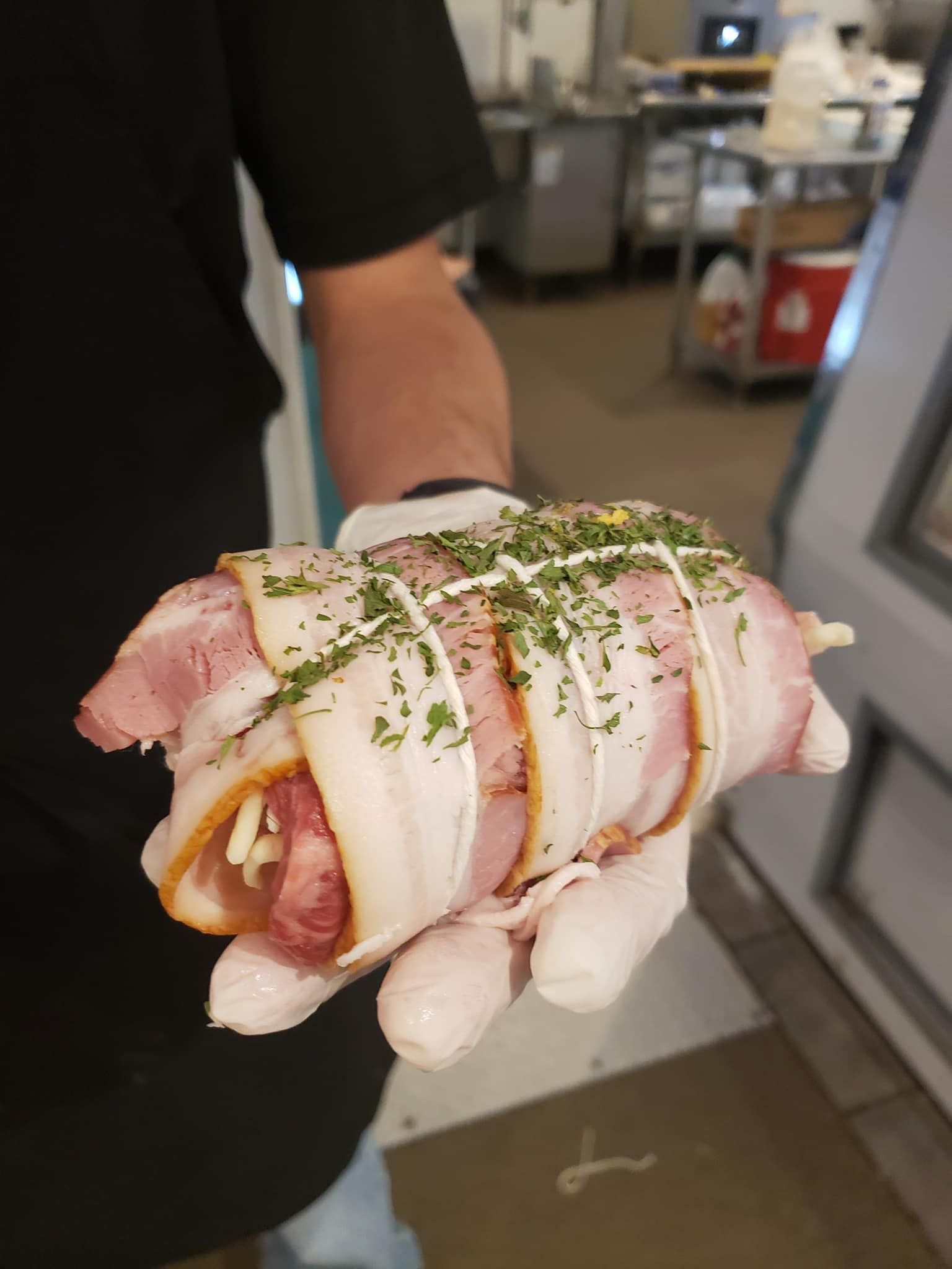 Person in white gloves holds a bacon-wrapped sausage, garnished with herbs and sauce, in a kitchen.