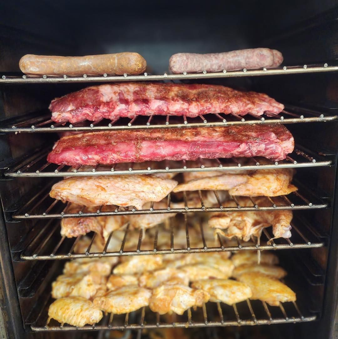 Smoker filled with ribs, sausage, chicken wings, and chicken, all seasoned and arranged on racks.