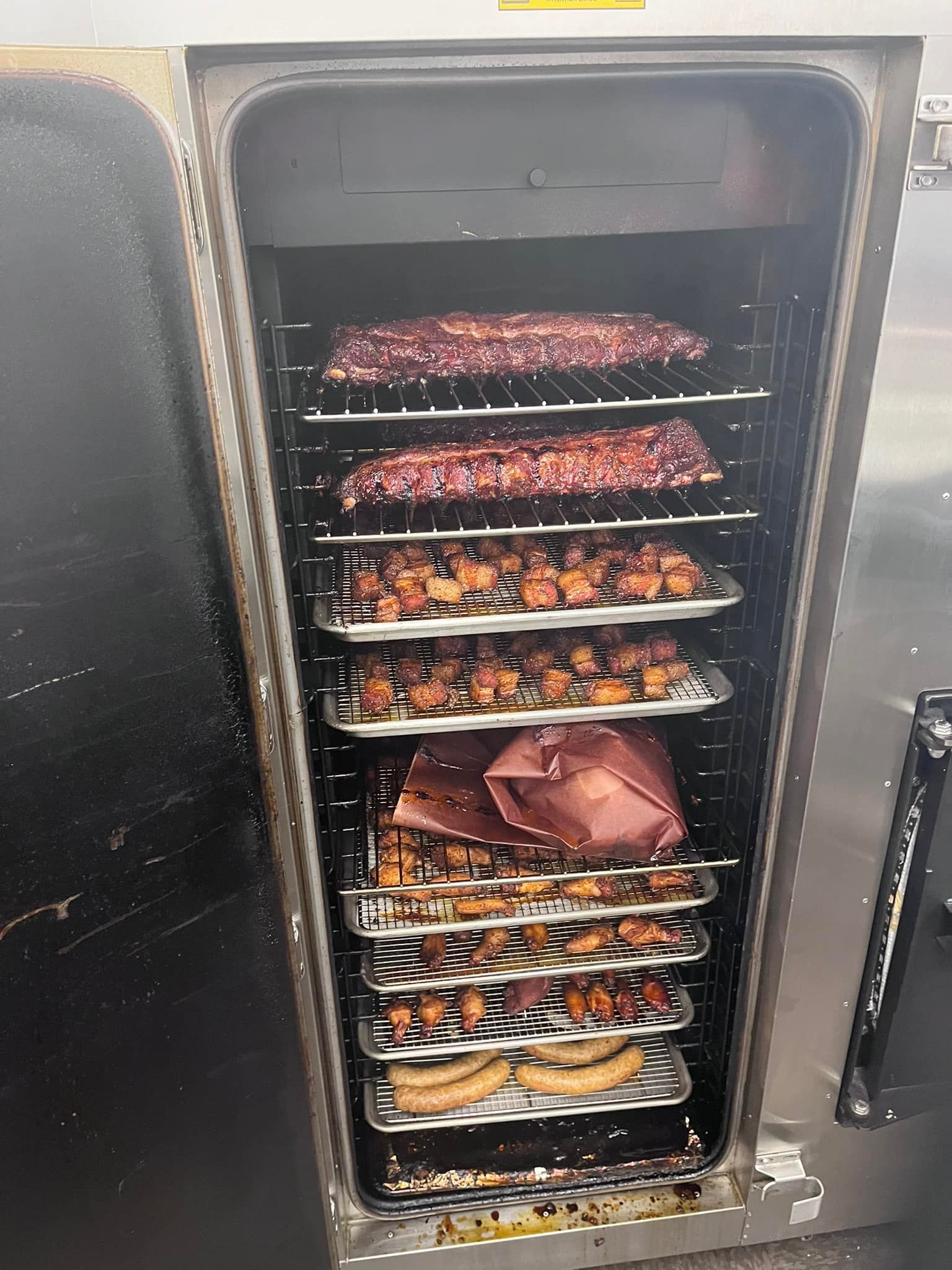 Smoker filled with various meats: ribs, pork belly, sausage, with open door.