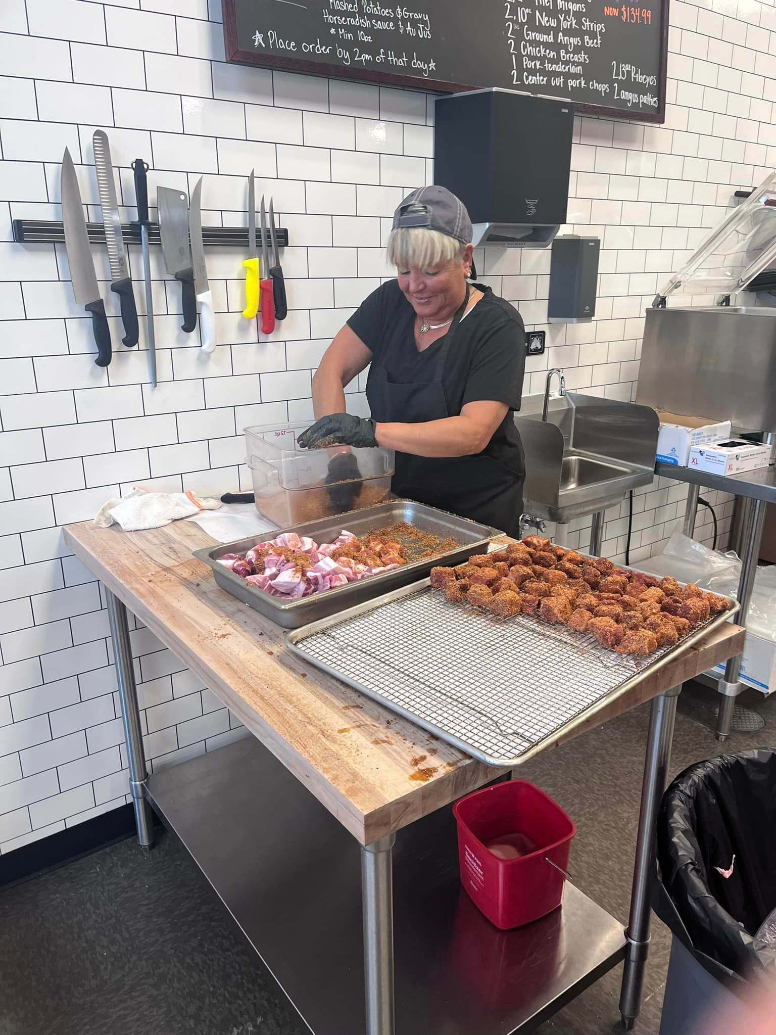 A woman wearing a cap and gloves prepares food behind a stainless steel table in a kitchen setting.