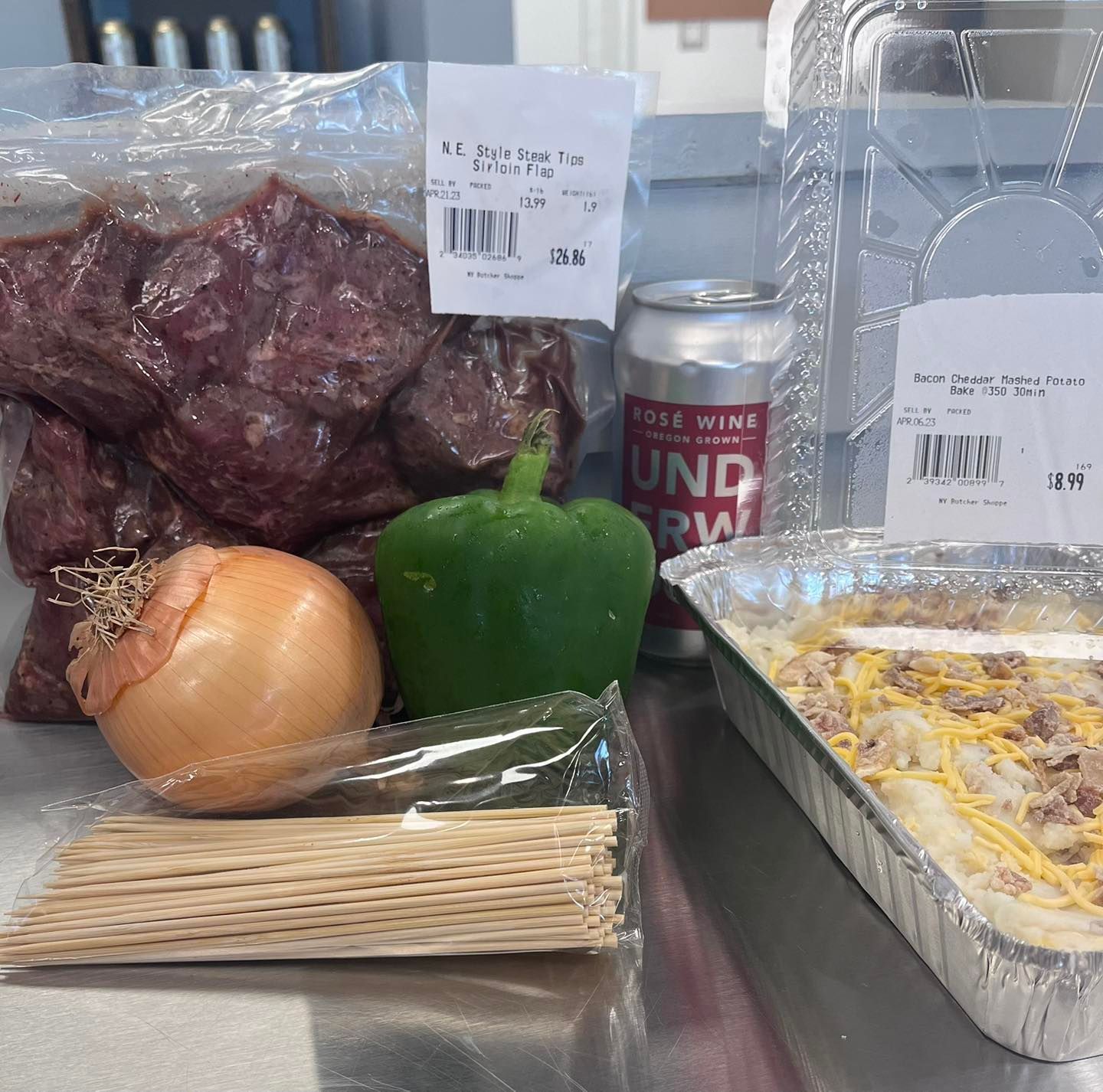 Ingredients for a meal including packaged meat, onion, green pepper, beer, and a casserole in a metal pan.