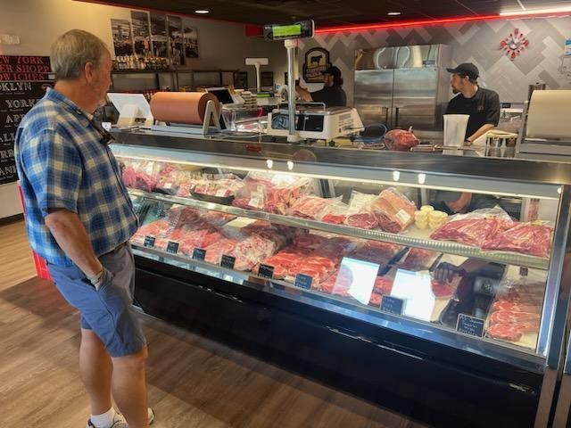 A man in a blue shirt looks at meat in a refrigerated display case at a butcher shop.