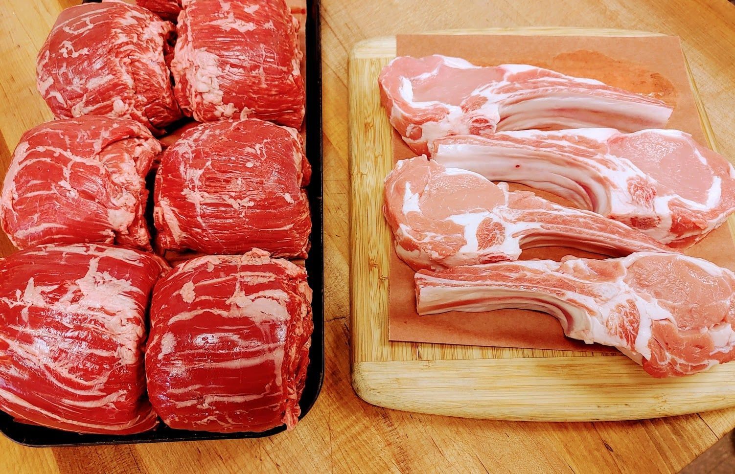 Raw cuts of red meat, including steaks and pork chops, on wooden cutting board.
