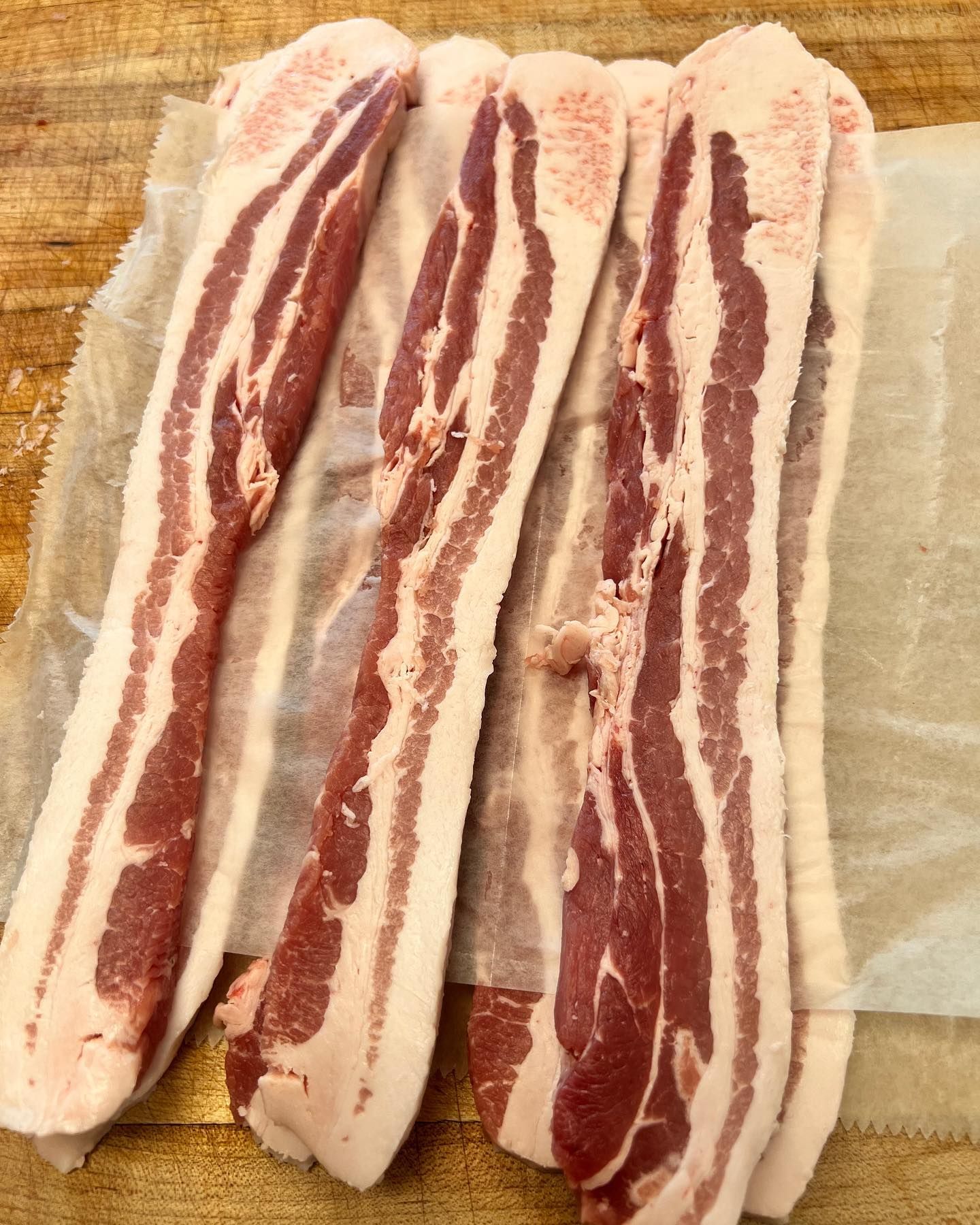 Four strips of raw bacon on parchment paper, on a wooden surface.