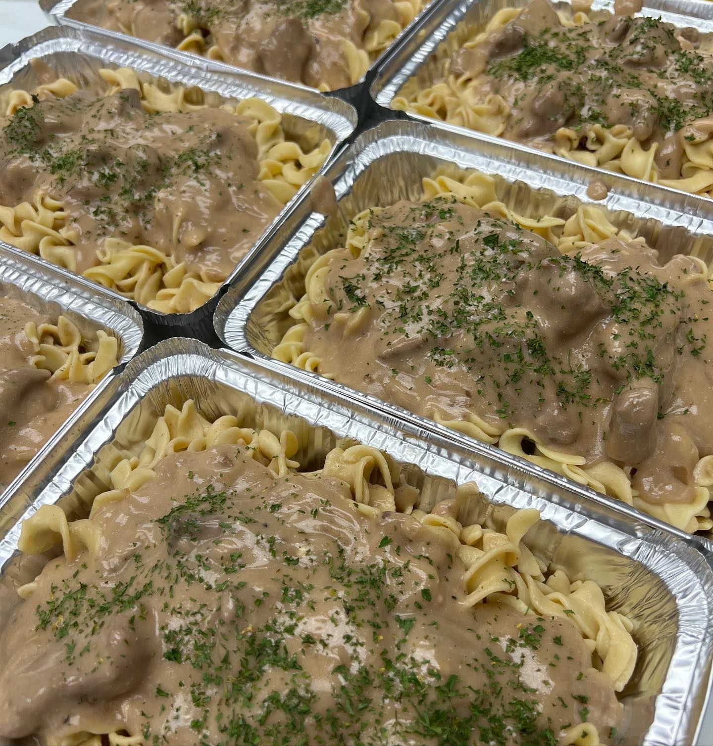 Several aluminum trays filled with egg noodles topped with a creamy gravy and parsley garnish.
