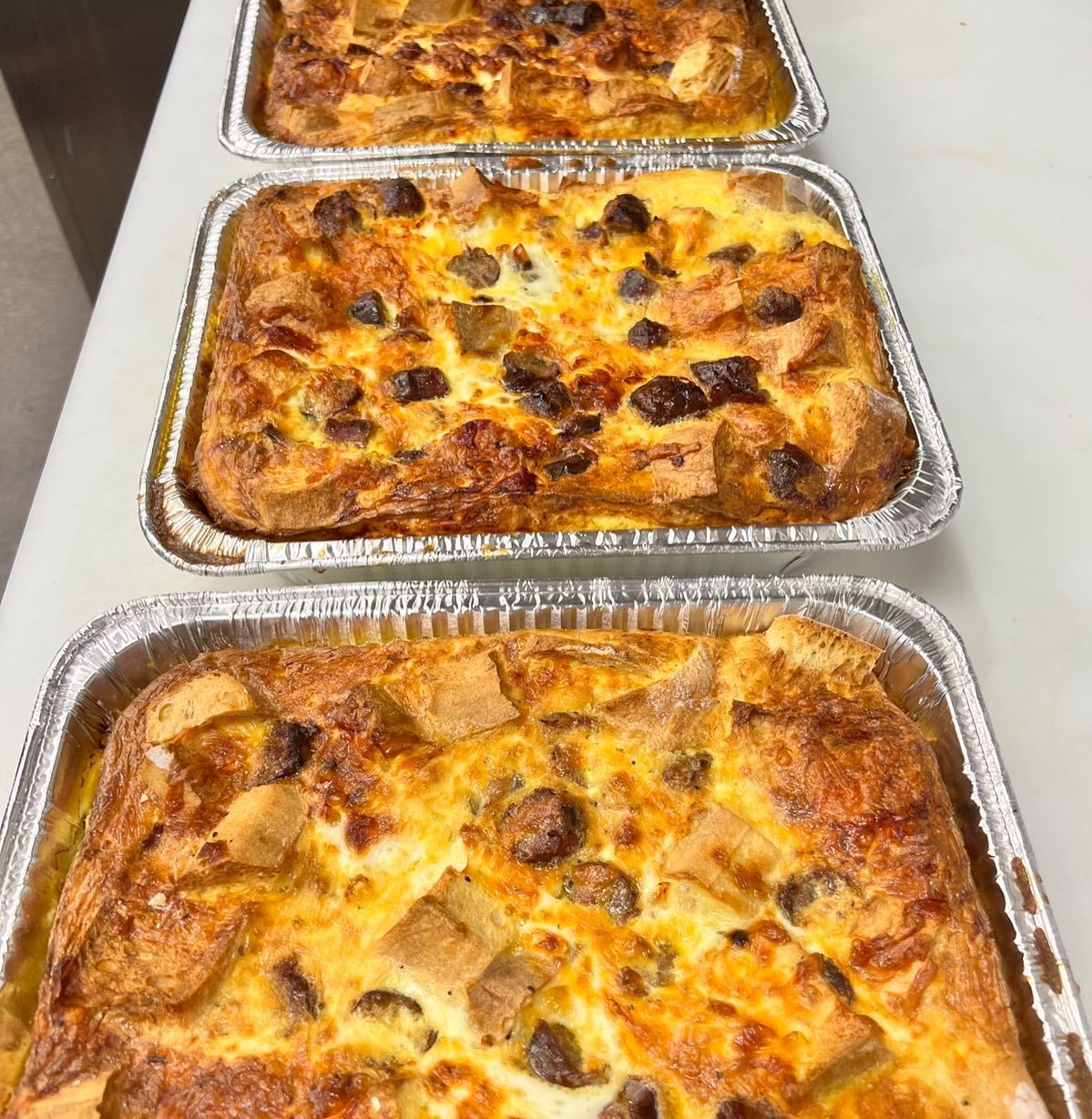 Three aluminum trays of baked casserole; golden-brown surface with visible bread cubes and dark pieces.