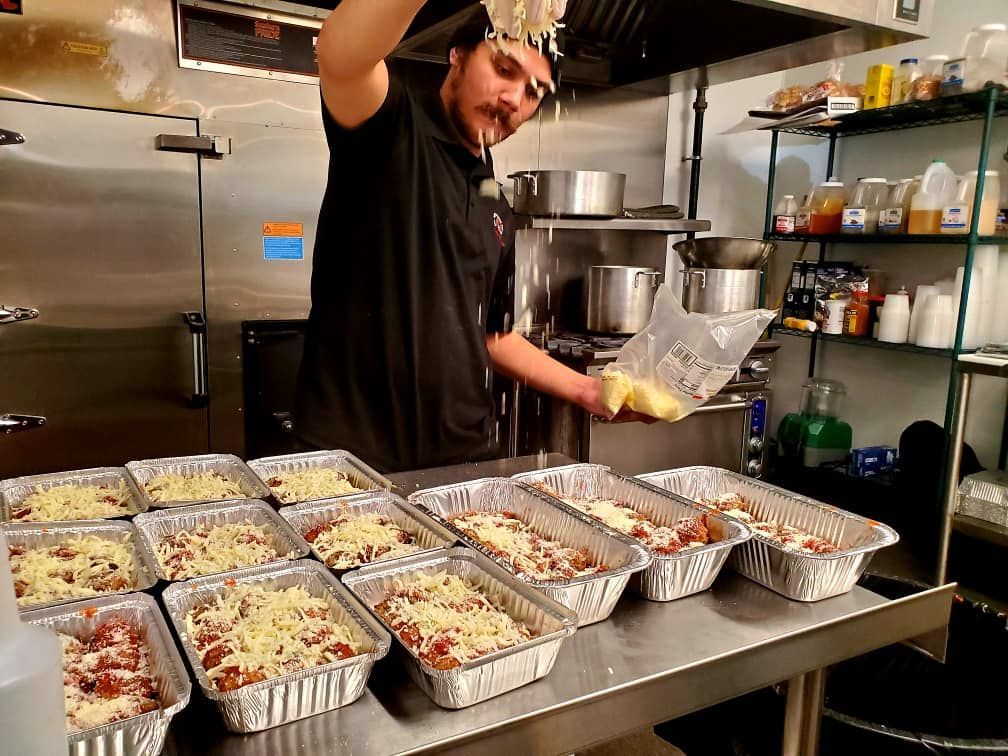 Chef topping lasagna in aluminum trays with cheese in a commercial kitchen.