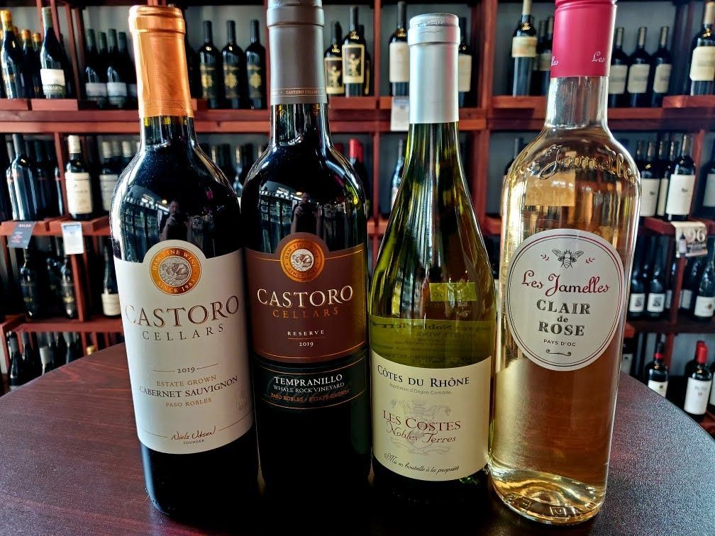 Four bottles of wine on a table in front of a shelf filled with wine bottles.