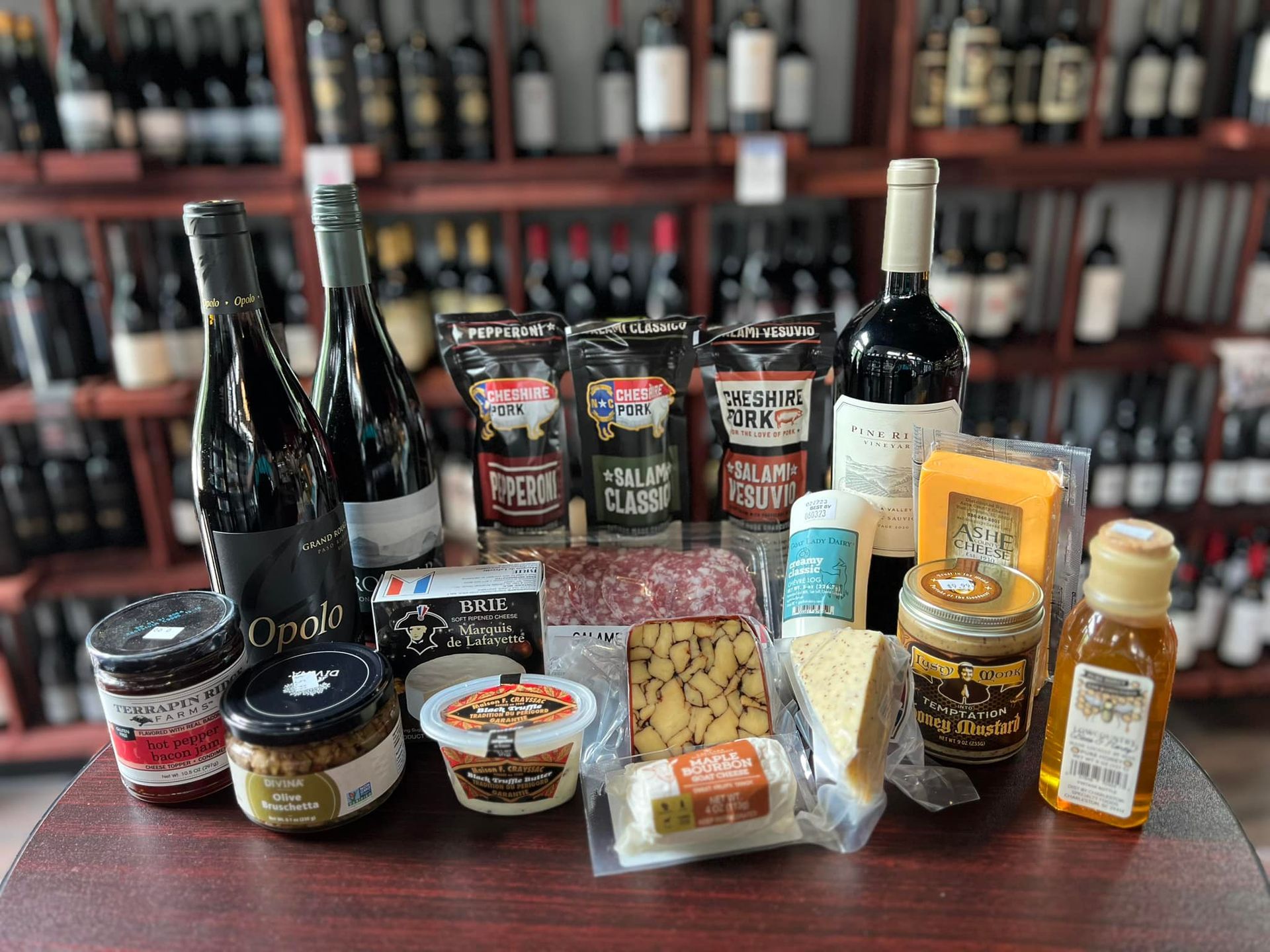 A display of wines, cheeses, meats, and preserves on a table in a store, surrounded by shelves of bottles.