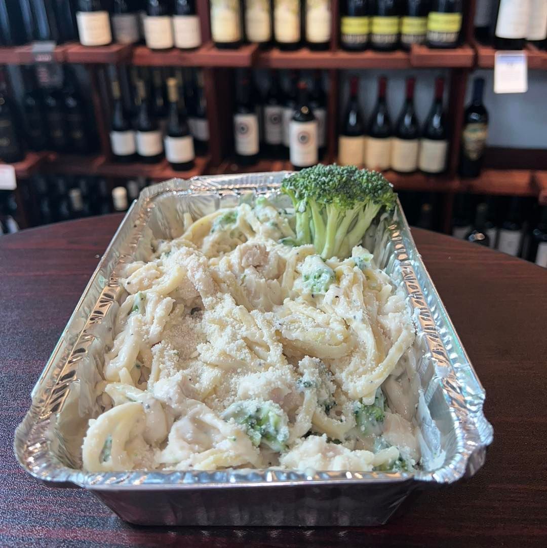 Pasta dish in foil container, broccoli, cream sauce, grated cheese, wine bottles in background.