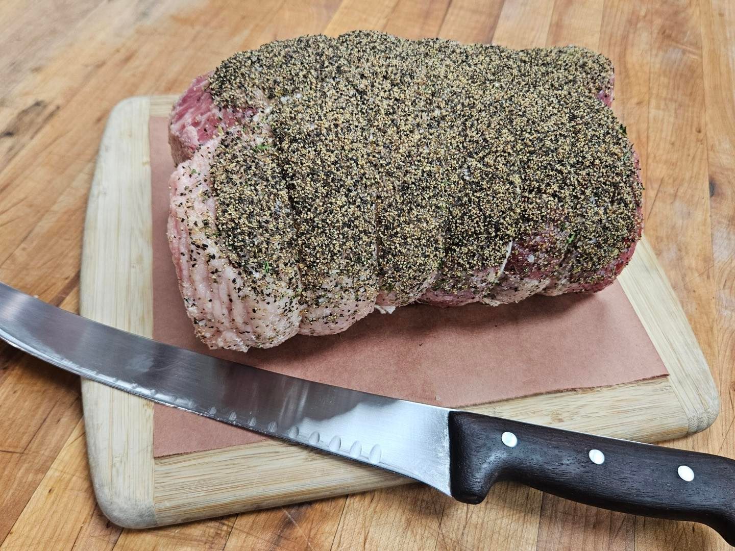 Raw beef roast seasoned with spices, resting on butcher paper and a wooden cutting board with a knife.