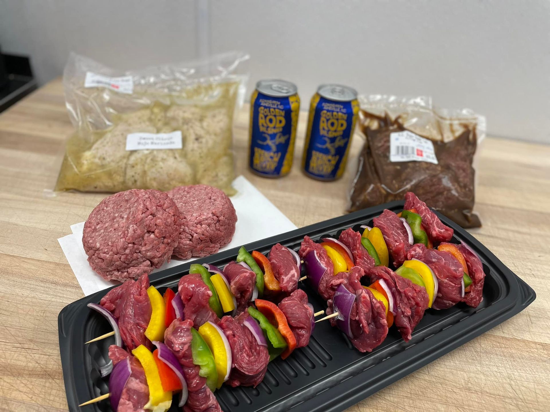 Meat skewers, ground meat, marinades, beer cans on a counter; ready to grill.
