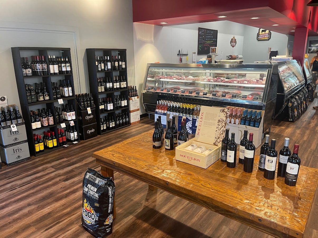 Wine shop interior: Wooden table with wine bottles, wine racks, deli counter with meats, and wood-paneled flooring.