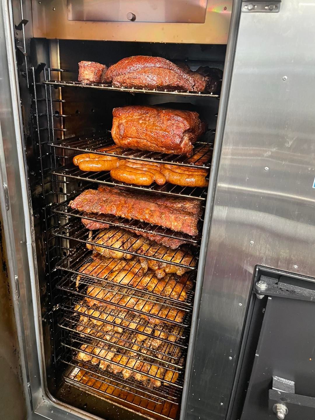 Smoker filled with various smoked meats including ribs, chicken, sausage, and pork, in a stainless steel setting.