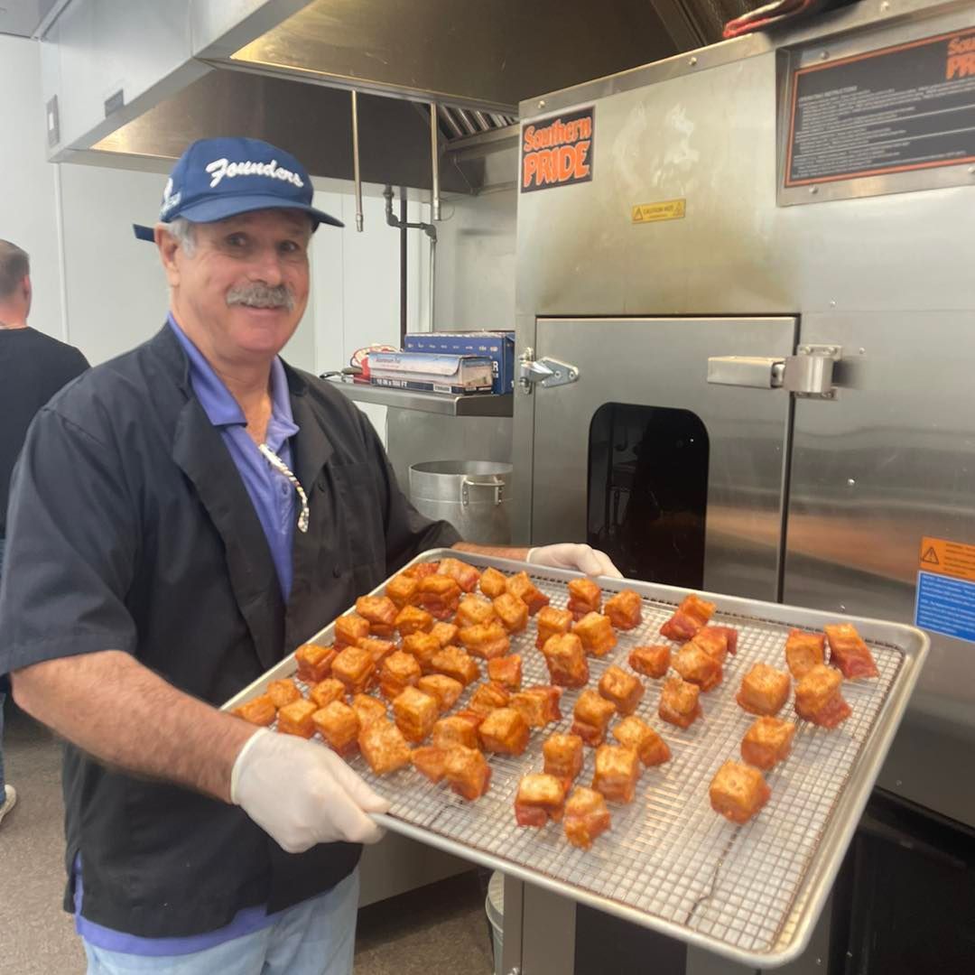 Man holding a tray of golden-brown fried food in a commercial kitchen. He's smiling, wearing a hat, and gloves.