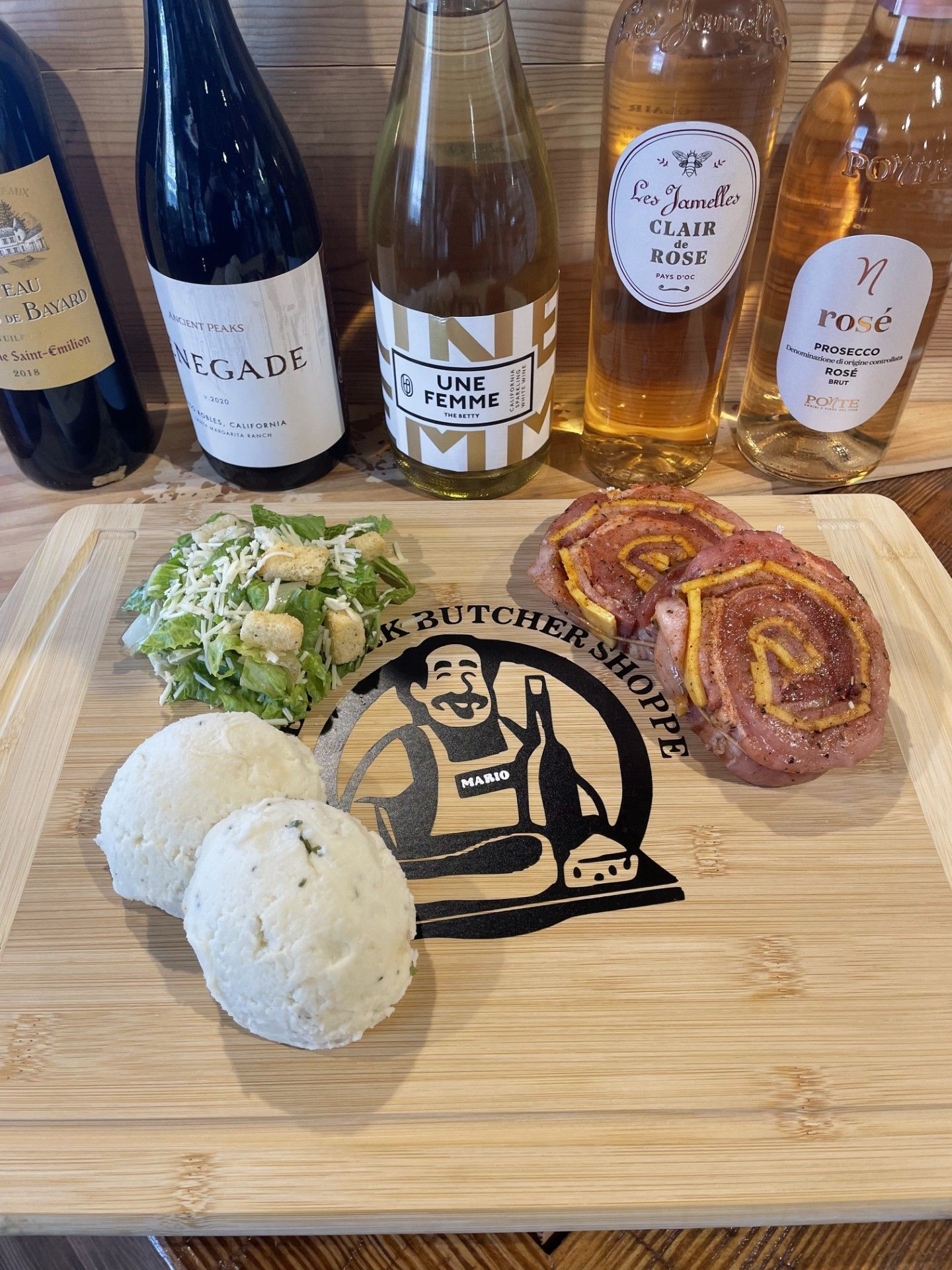 Wooden cutting board with food and wine bottles. Featuring salami rolls, cheese balls, and salad in front of various wine bottles.