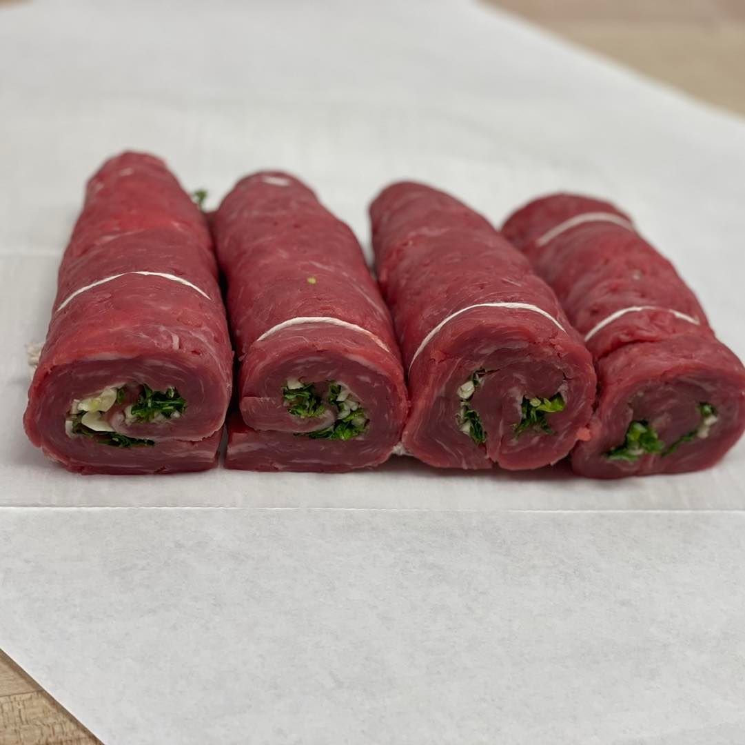 Four beef braciole rolls on white paper. Meat is red, filling is green, string tied around.
