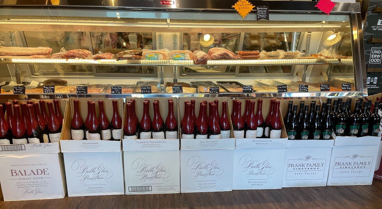 Bottles of wine displayed in white boxes in front of a deli counter, with meats and pastries visible.