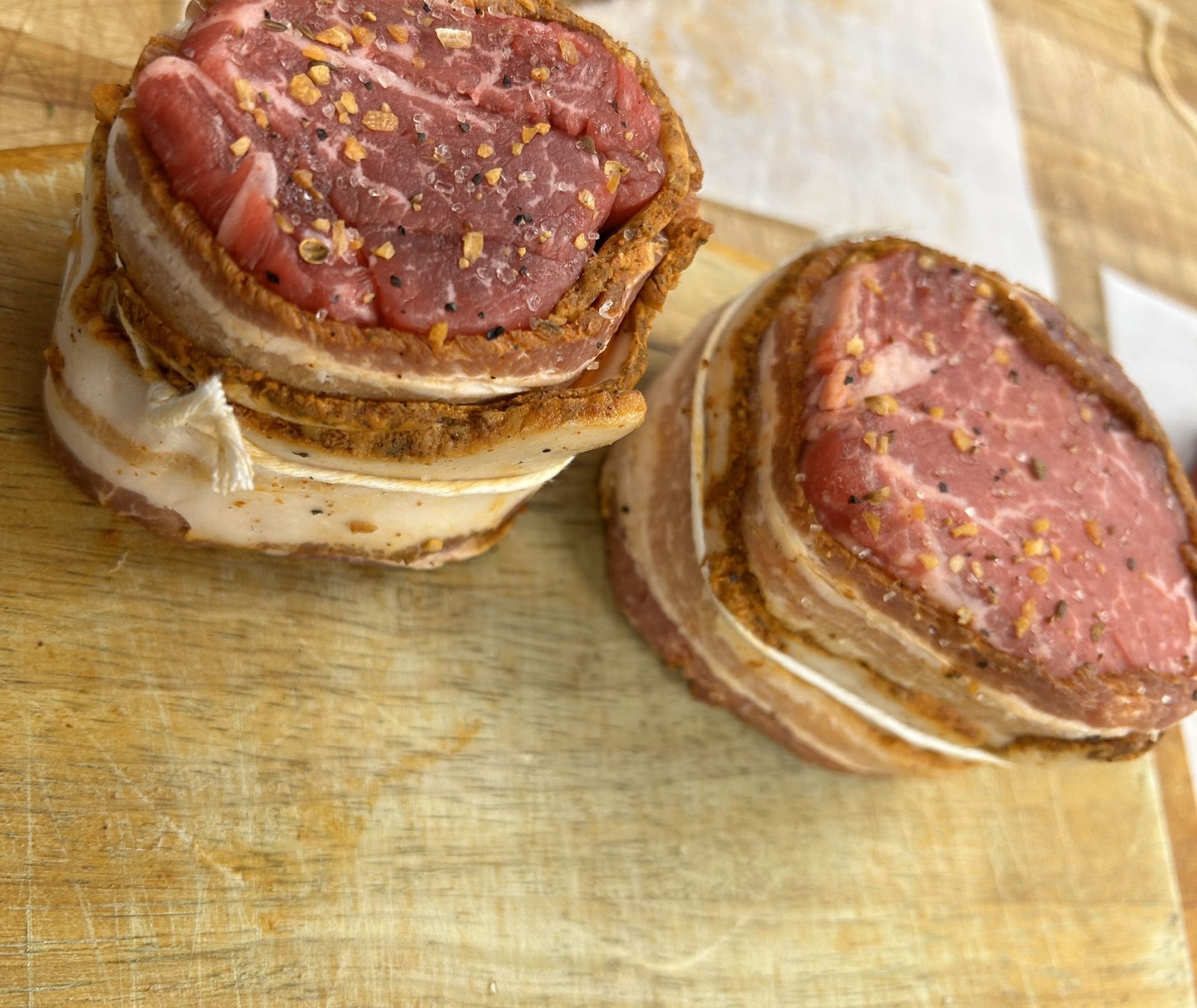 Two bacon-wrapped, seasoned filet mignon steaks on a wooden cutting board.