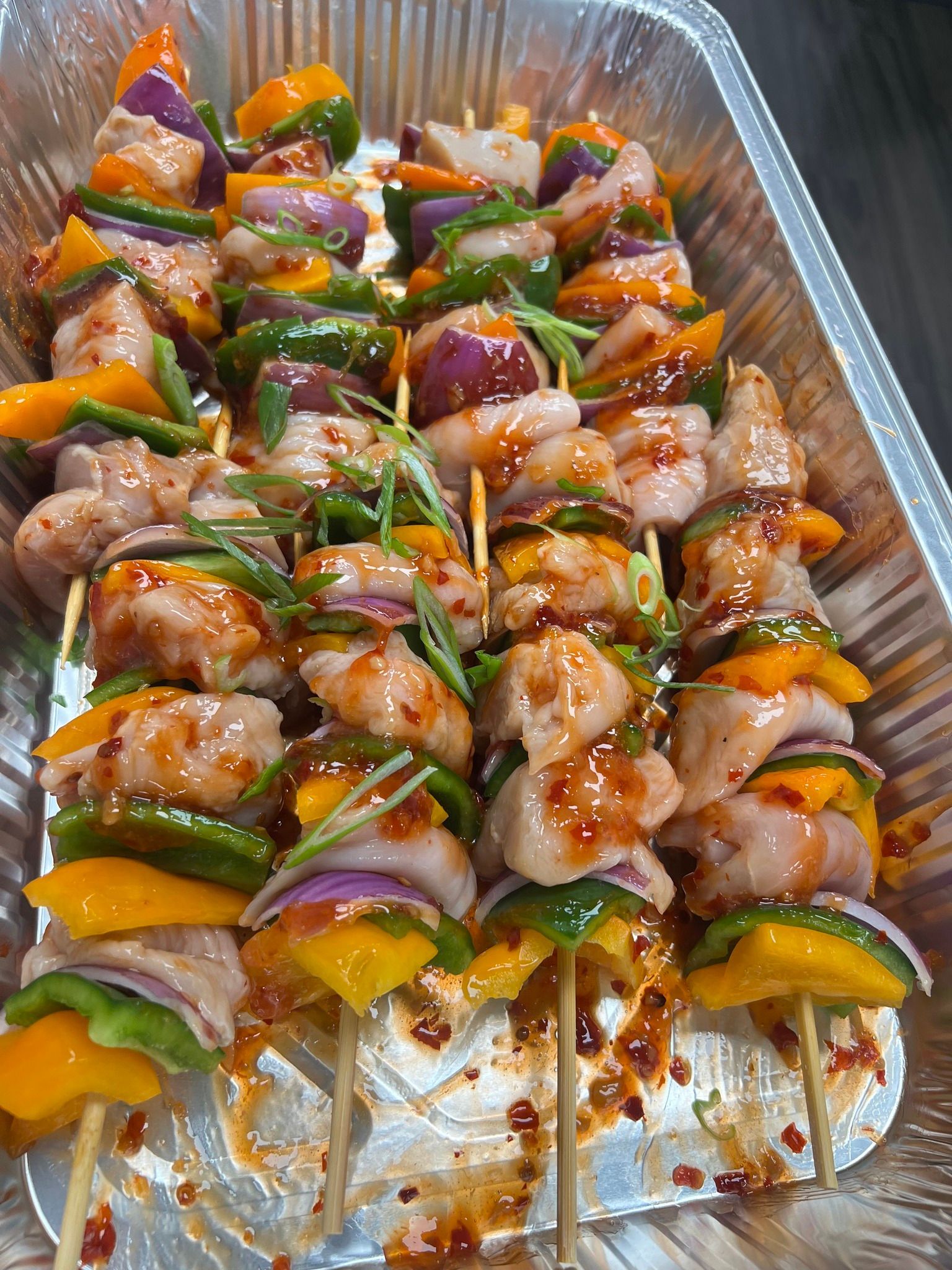 Chicken and vegetable skewers in a foil pan, seasoned with sauce and garnished with chives.