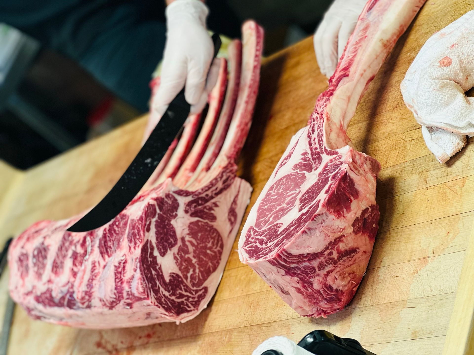 A butcher cutting a large, raw tomahawk steak on a wooden block, wearing gloves.