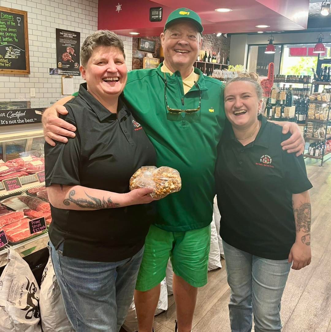 Three smiling people in a store; two women in black shirts flank a man in green. One woman holds a loaf.