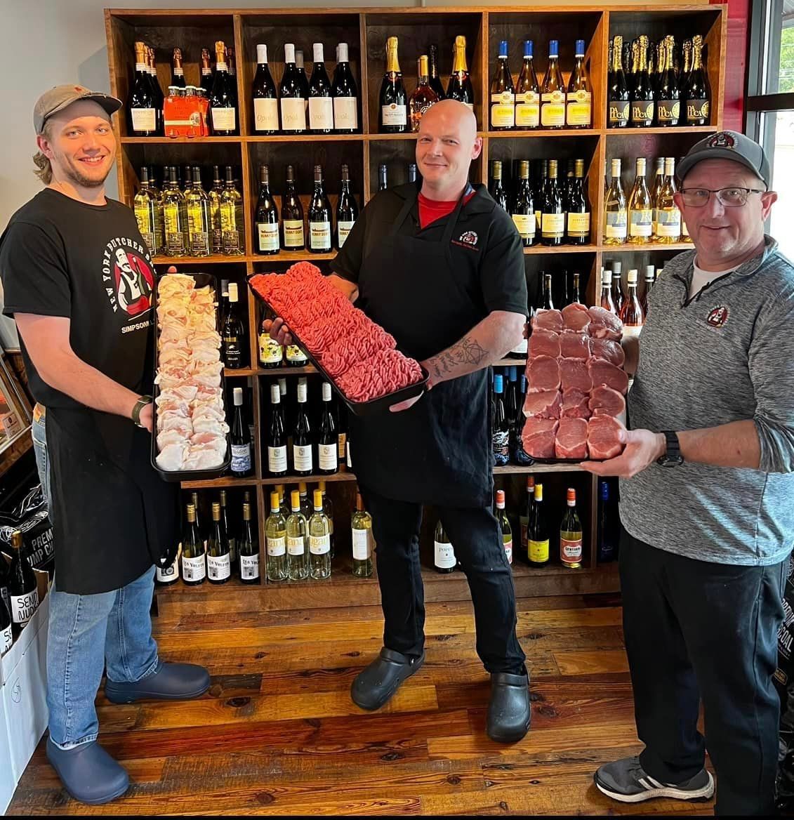 Three men in a shop holding trays of meat with wine bottles in the background.