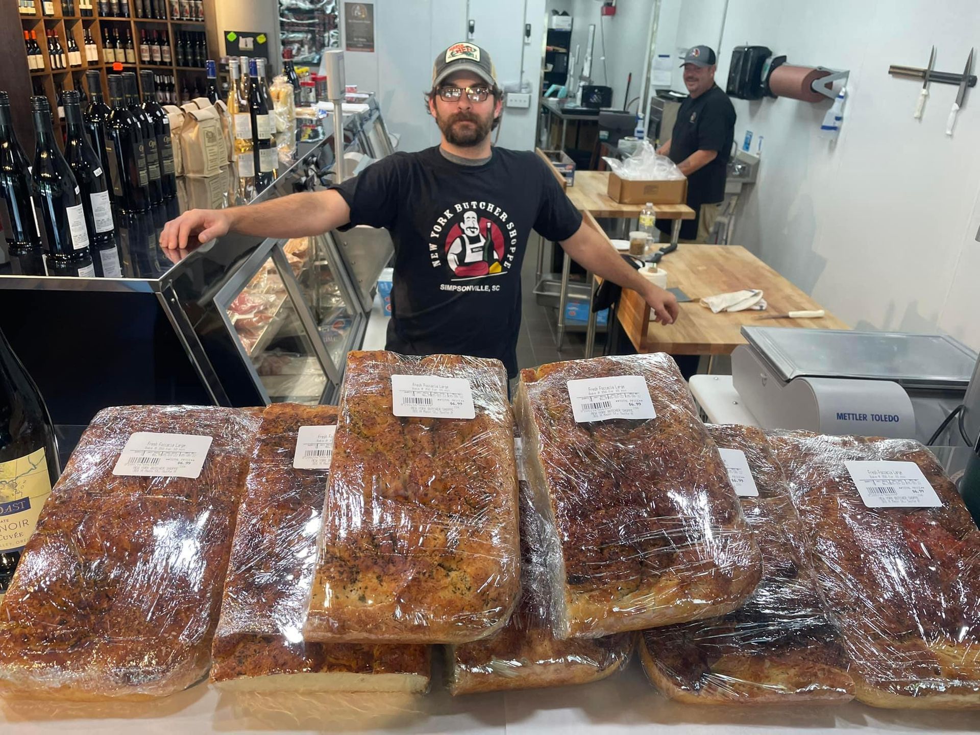 Man stands behind a counter with packaged focaccia bread. A wine shop interior.