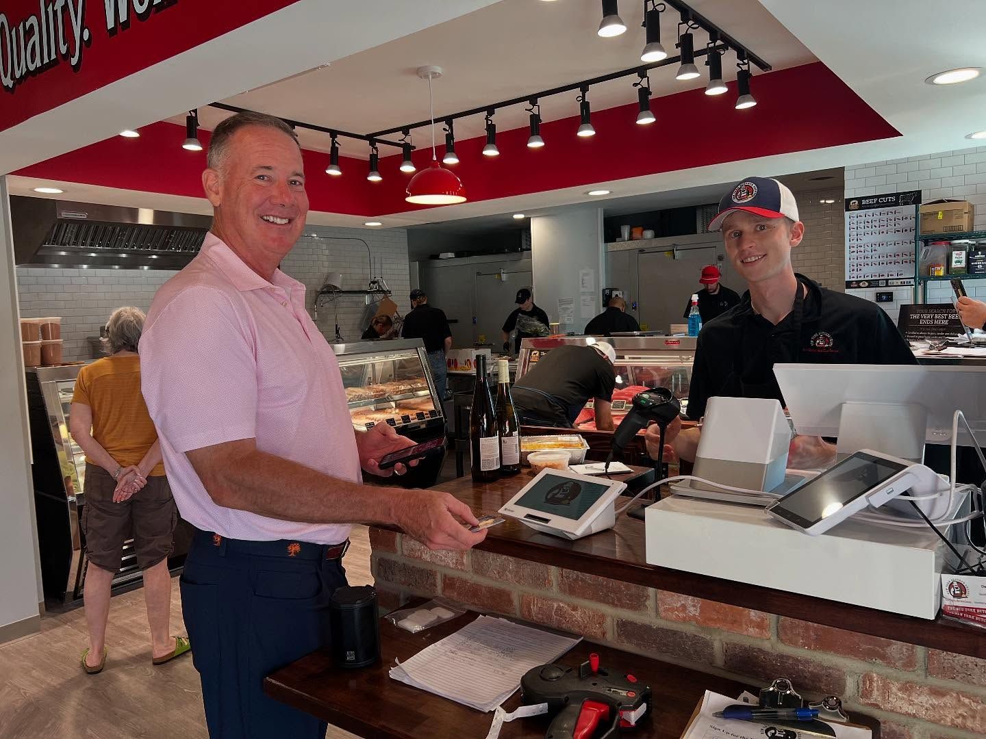 Man paying at butcher shop counter; worker smiles. Shop has red accents, brick, and a deli case.