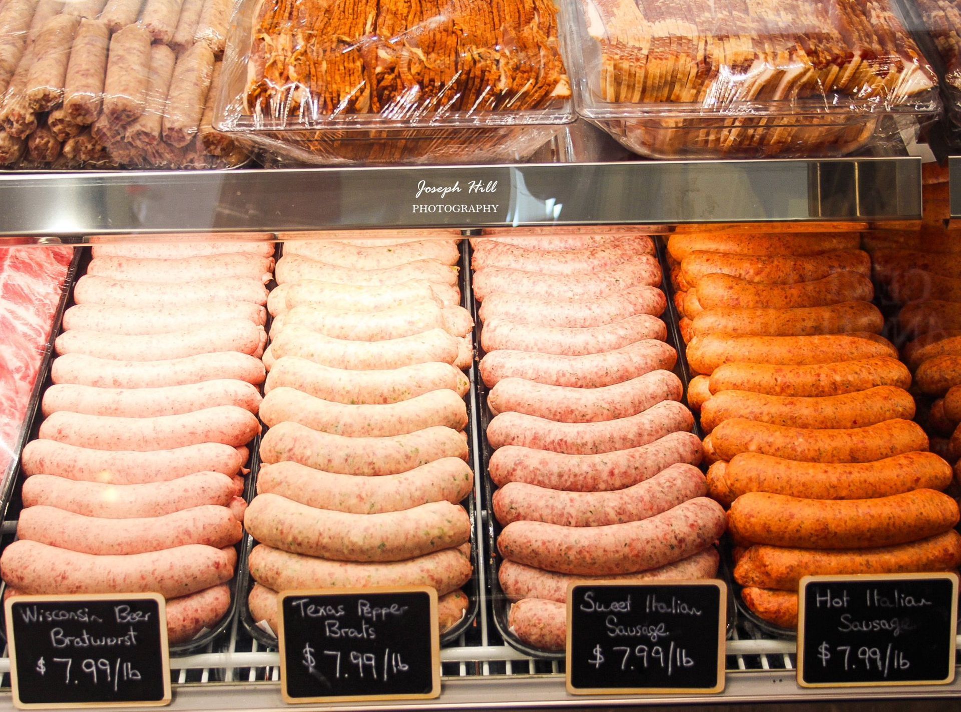 Rows of various sausages in a refrigerated display case with price labels, butcher shop.