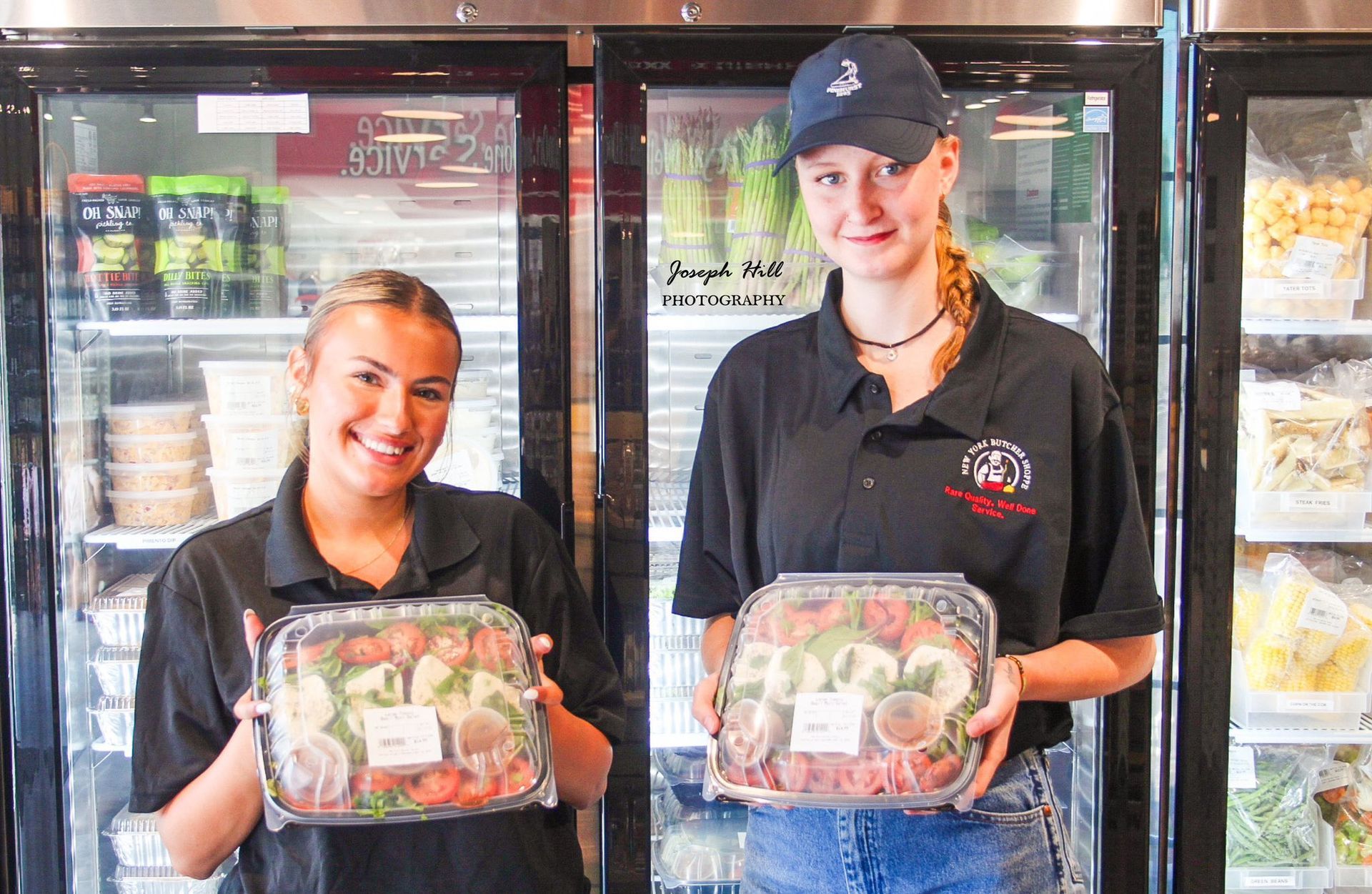 Two women smiling, holding prepared food trays in front of refrigerated display cases.